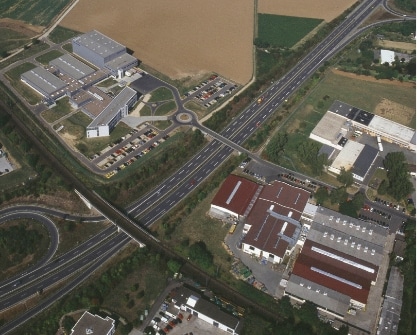 Luftaufnahme eines großen Industriekomplexes mit mehreren Gebäuden und Anlagen, umgeben von Grünflächen und Straßen.